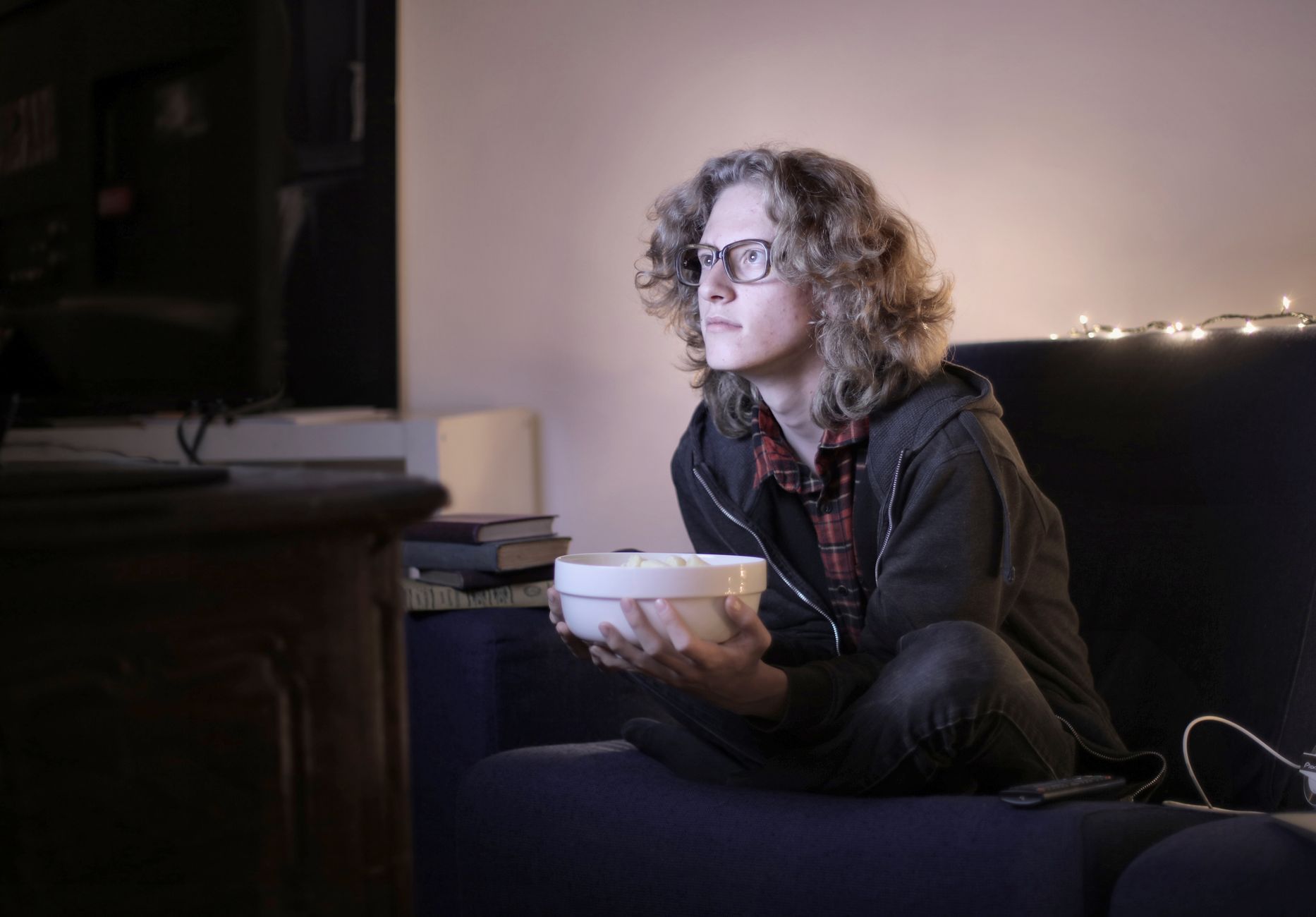 Concentrated male teenage with long hair in casual wear and eyeglasses sitting on couch with bowl of popcorn and watching interesting movie 