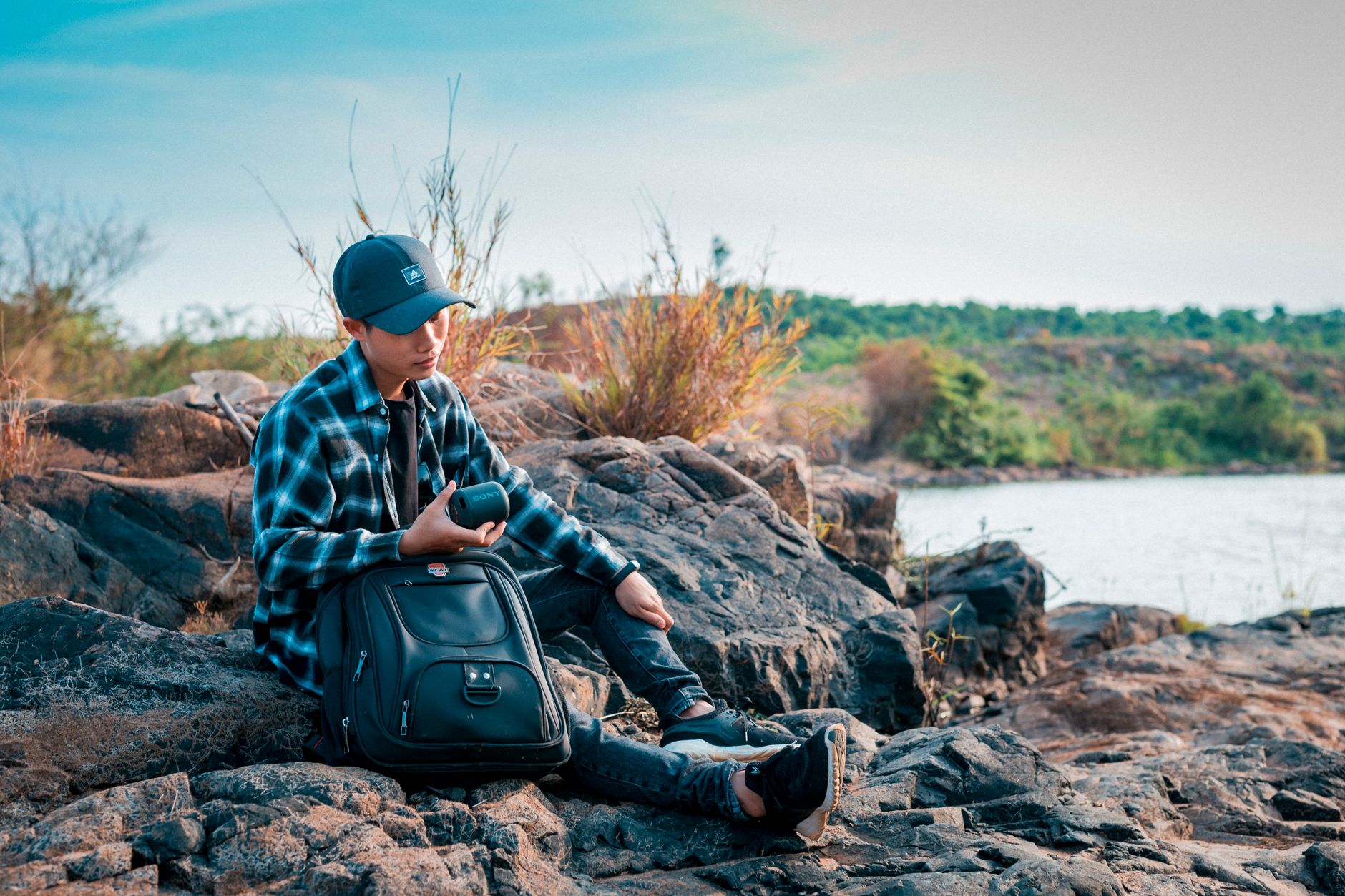 Unrecognizable young traveler with rucksack listening to music from portable loudspeaker while sitting on rocky shore near river during vaca