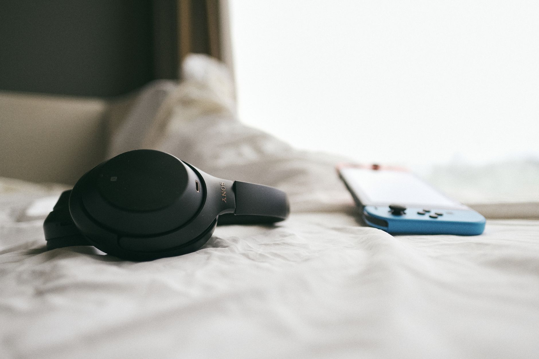 Close-up of a black headset and a gaming console resting on a soft, white bedspread in a calm, indoor setting.