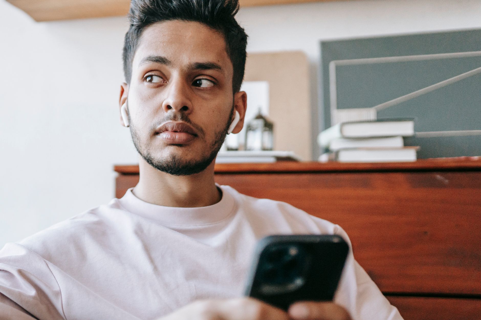 Serious Indian man listening to music in wireless earbuds looking away while texting message on cellphone in light room against table with b