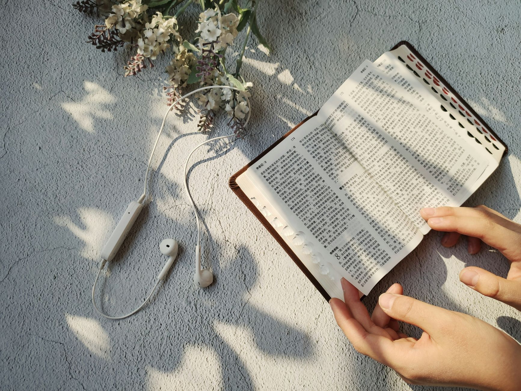 Serene scene of hands reading a book with flowers and earbuds on a sunny day.