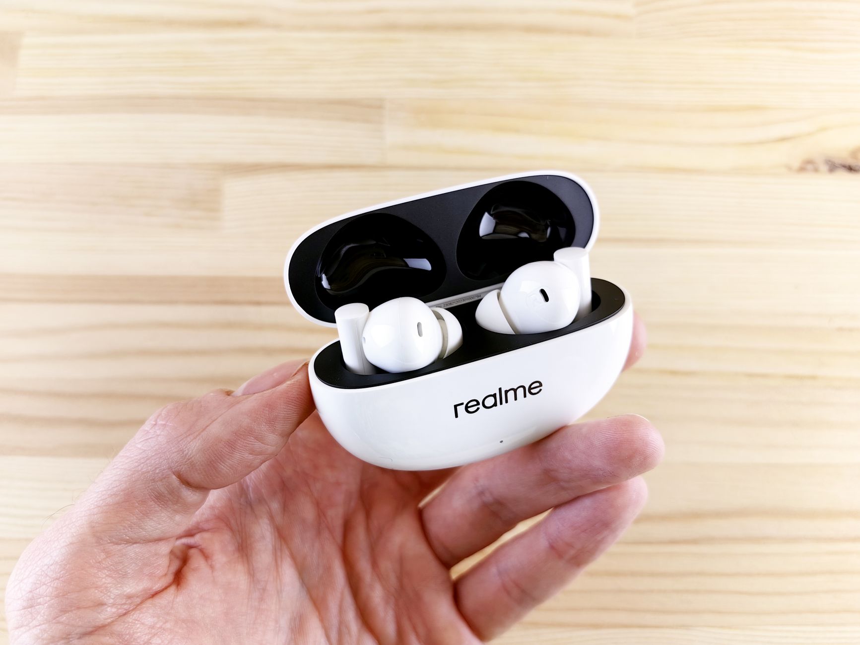White wireless earbuds in open charging case held by hand on a wooden surface.