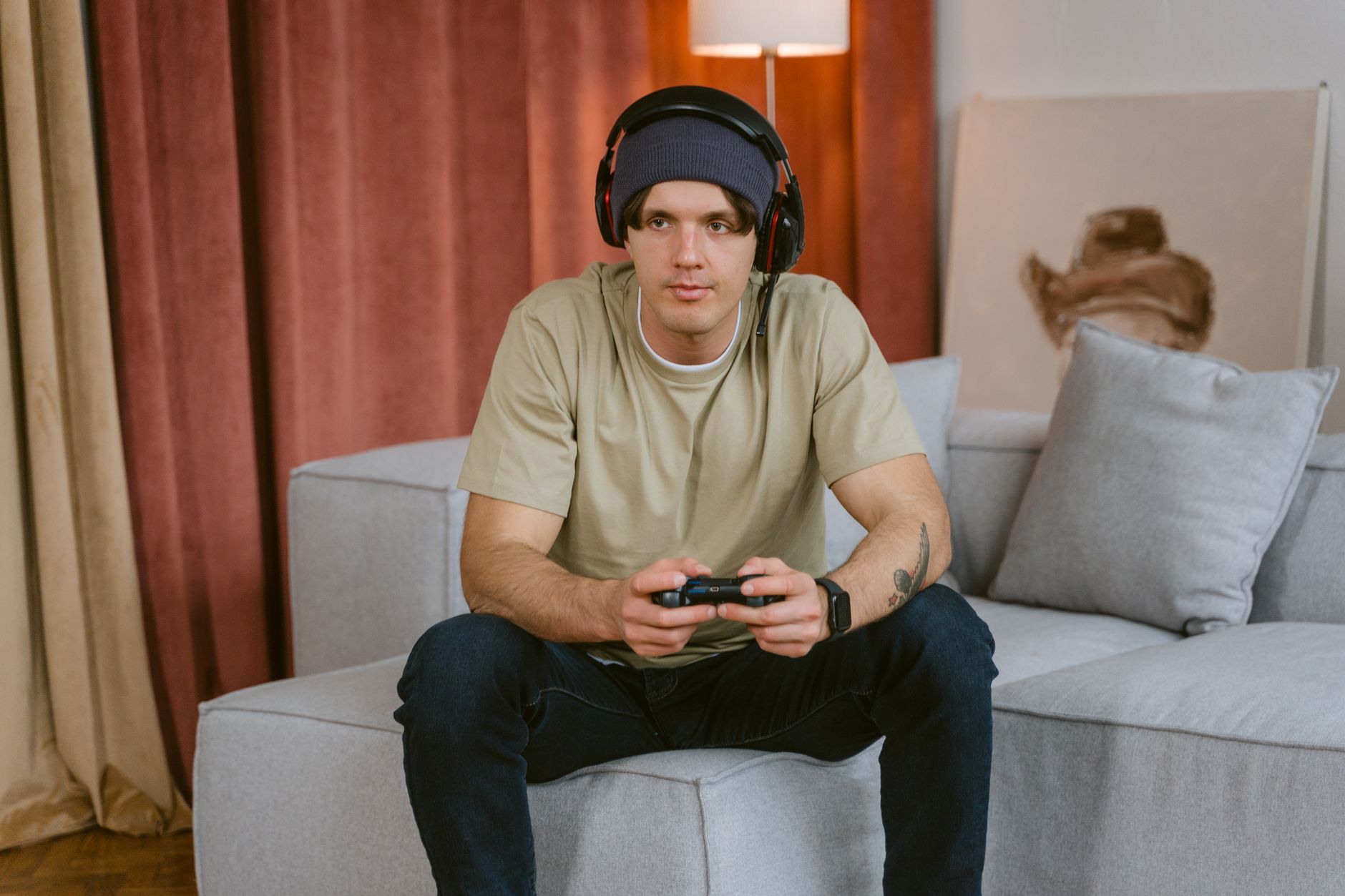 A young man wearing a beanie and wireless headphones, playing video games on a couch.