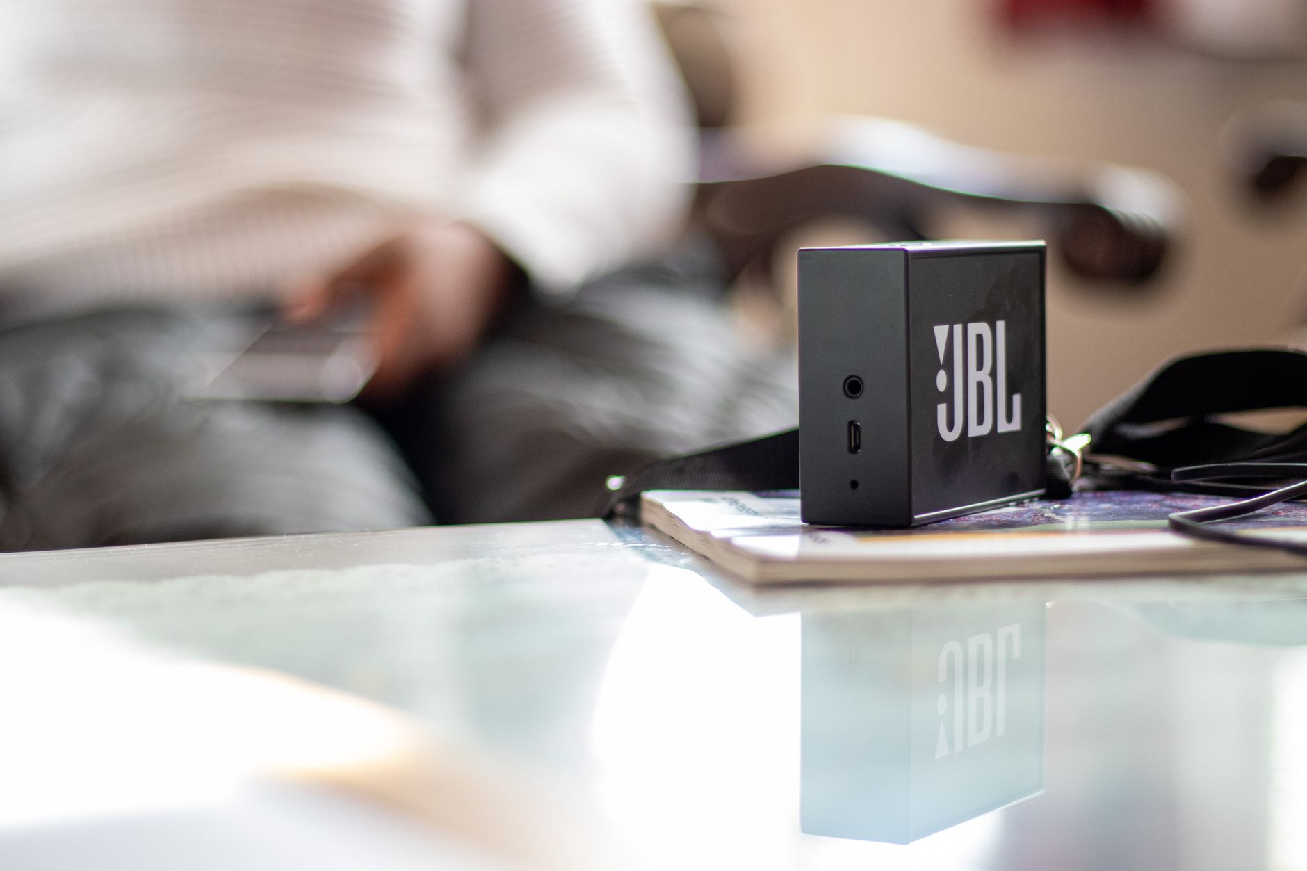 A close-up of a JBL speaker on a reflective table indoors, with a blurred background and natural light