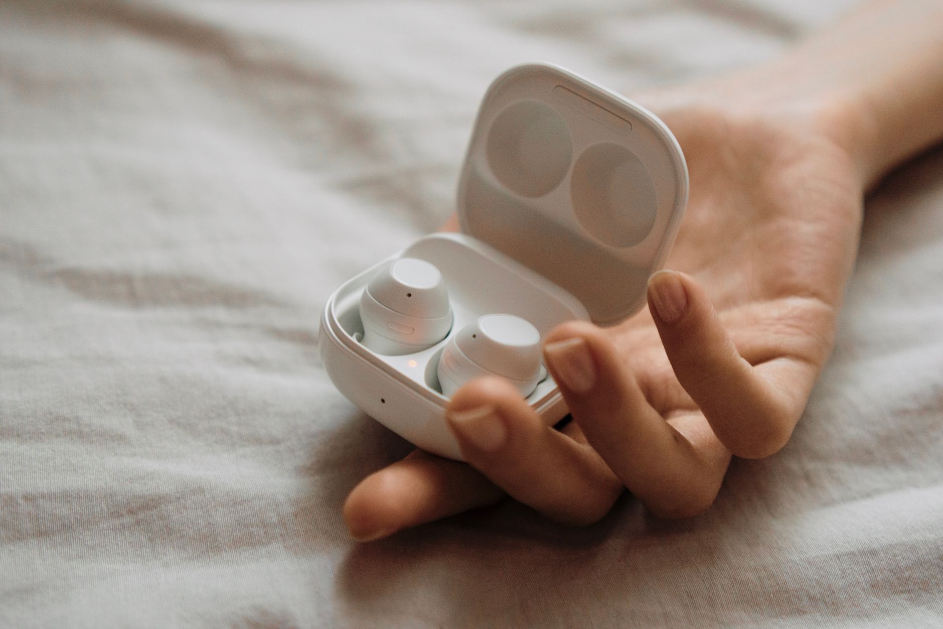 Close-up of a hand holding white wireless earbuds in an open case, resting on a soft surface.