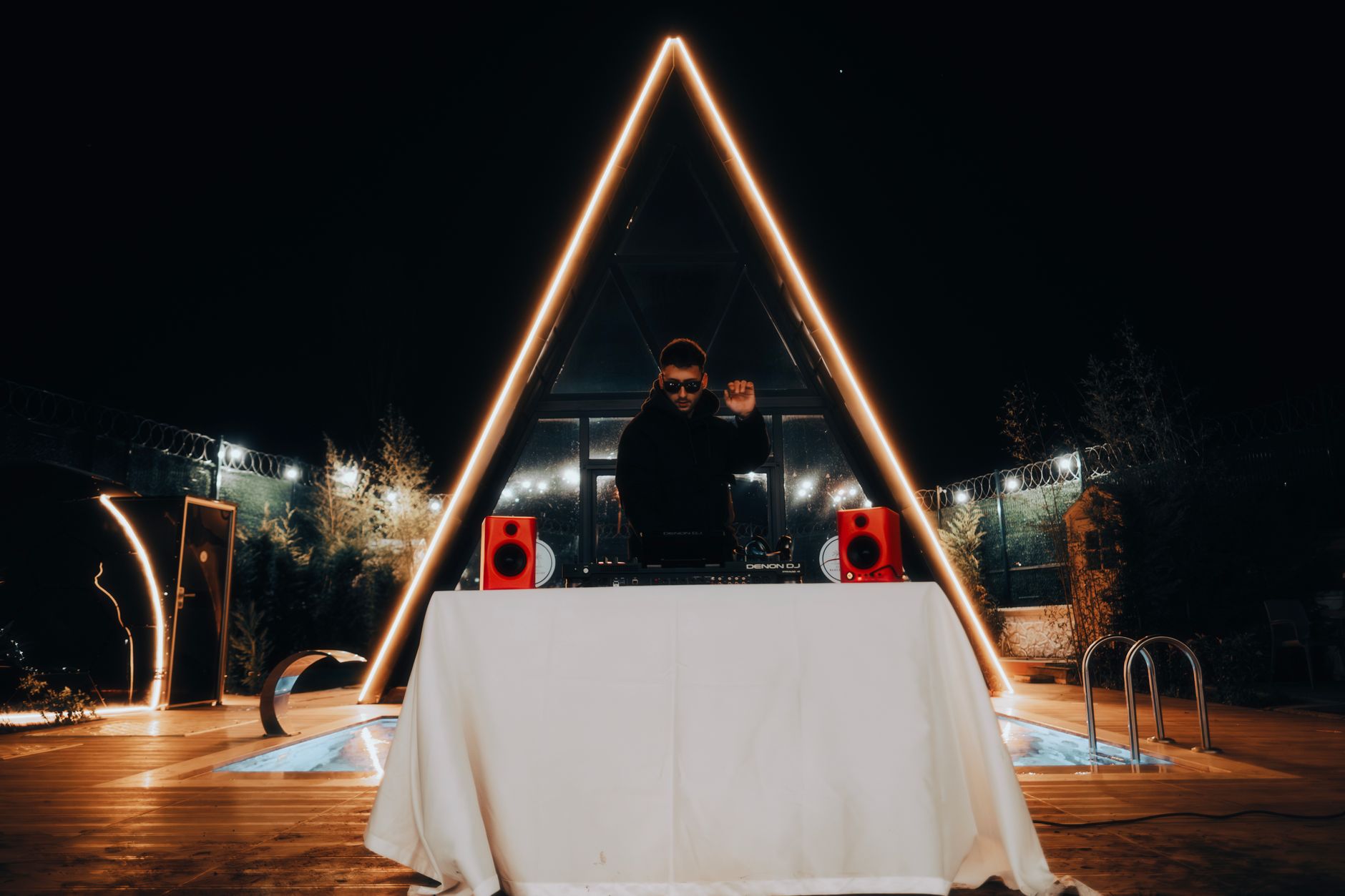 A DJ performing at an outdoor pool party at night, with vibrant lights and sound equipment.