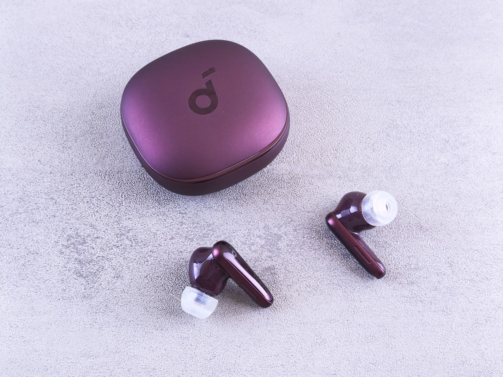 Modern purple wireless earbuds and charging case on a textured concrete surface.