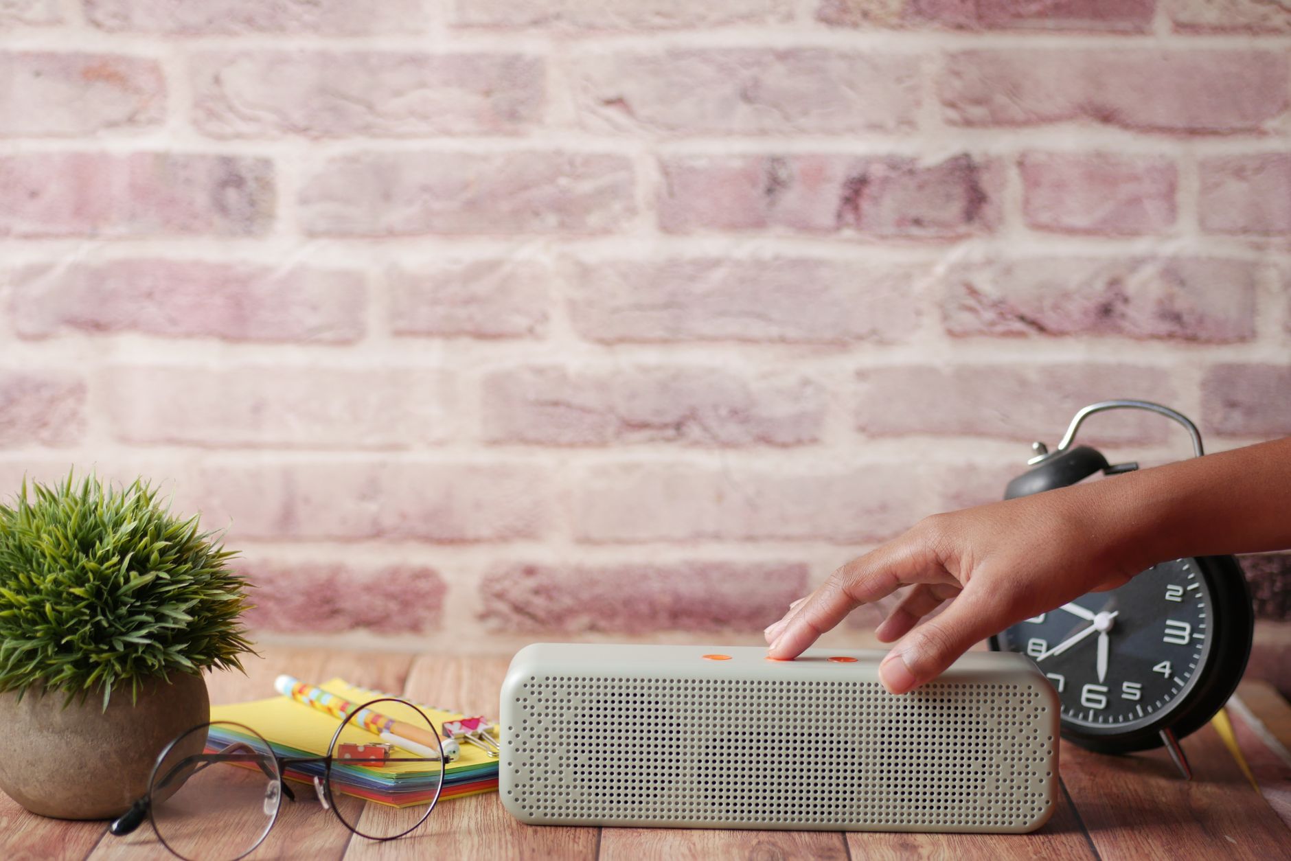 Close-up of a hand touching a wireless speaker on a desk with various office items.