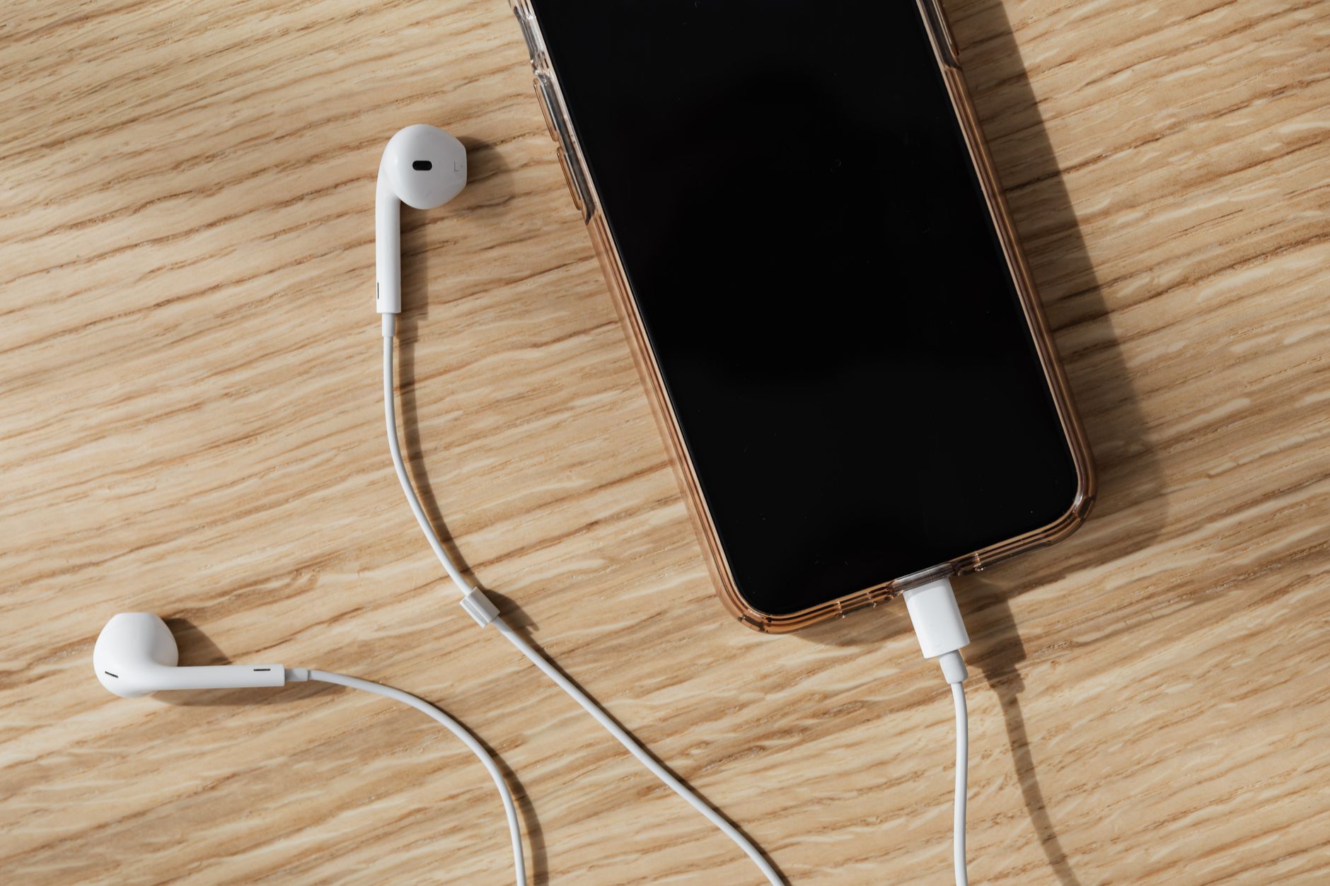 From above composition of white wired earphones and modern mobile phone with blank screen and connected charging cable placed on wooden surf