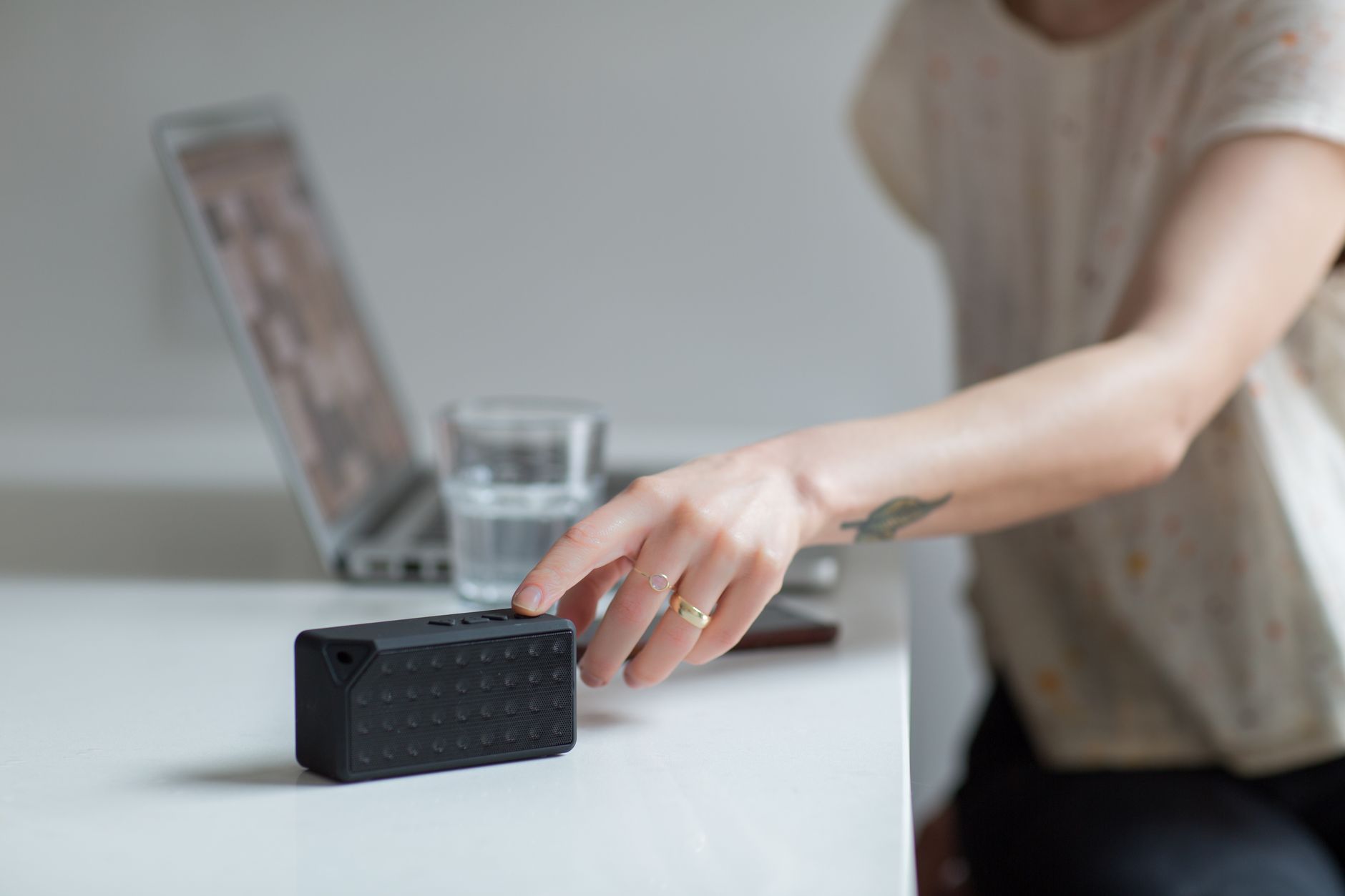 Close-up of a hand reaching for a wireless speaker on a desk beside a laptop.